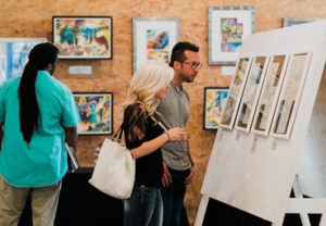 A man and woman standing and looking at artwork in a gallery. Another man with his back turned, looking at artwork.