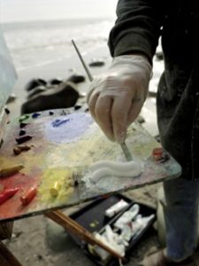 Gloved hand with paintbrush mixing white paint on a paint pallet.